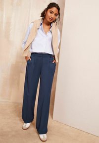 Helles blaues Hemd, cremefarbener Pullover über den Schultern, marineblaue weite Hose und weiße Sneakers mit braunen Akzenten.
