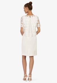Spitzenoberteil mit kurzen Ärmeln und floralem Design über einem schlichten weißen Kleid, das einen Schlitz im Rücken und einen geraden Schnitt aufweist, kombiniert mit Riemchenschuhen.