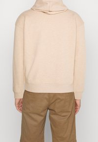 Felpa beige con una texture morbida, cappuccio ampio e vestibilità rilassata. Presenta polsini e orlo a costine, abbinata a pantaloncini marroni.