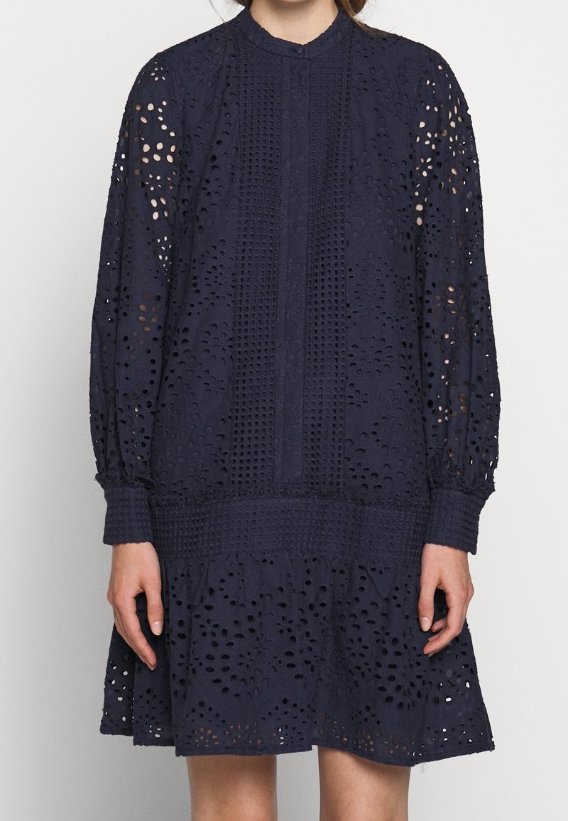 Robe bleu marine en dentelle avec découpes florales, manches longues, col montant et jupe à volants avec des accents texturés.