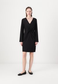 Mustern, pikse varrukatega must kleit V-kaelusega ja sõlmega vöökohal. Omab krohvitud siluetti ja põlvepikkust hemmi. Kandmiseks madala musta jalatsiga.