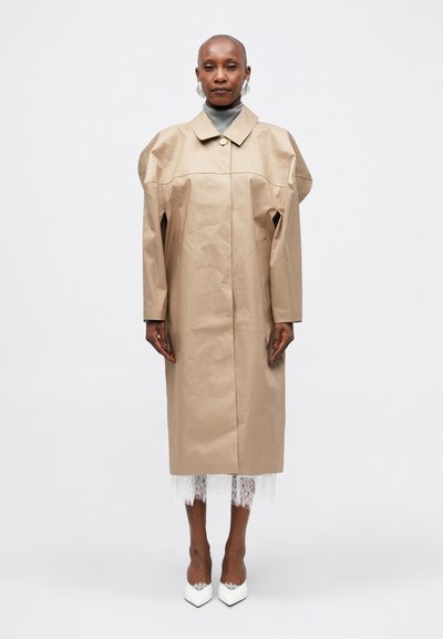 Manteau trench oversized beige avec un col, fermeture à boutons et un tissu texturé unique. Des chaussures blanches et un détail en dentelle sont visibles.