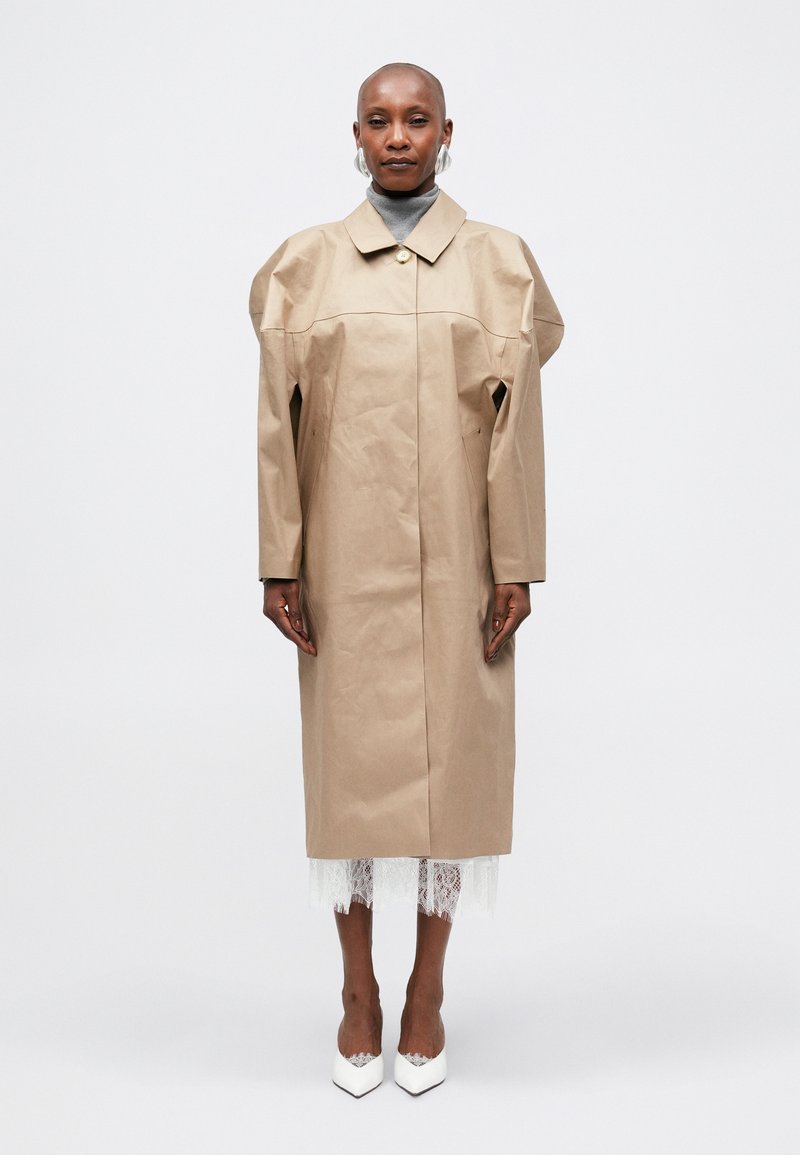 Béžový oversized trench coat s límečkem, zapínáním na knoflíky a unikátní texturovanou tkaninou. Bílé boty a detail krajky jsou viditelné.