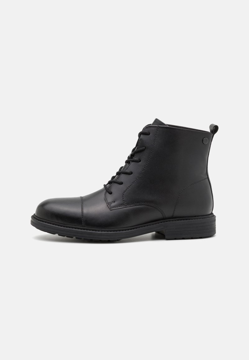 Bottine noire en cuir avec lacets à l’avant, bout rond, petit talon empilé et languette à l’arrière sur un fond clair uni.