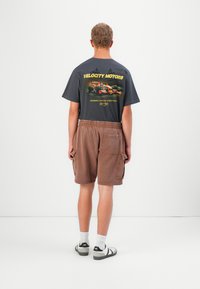 Mand iført sort grafisk racer T-shirt, brune cargo shorts, hvide sokker og hvide sneakers, står vendt mod en simpel hvid væg.