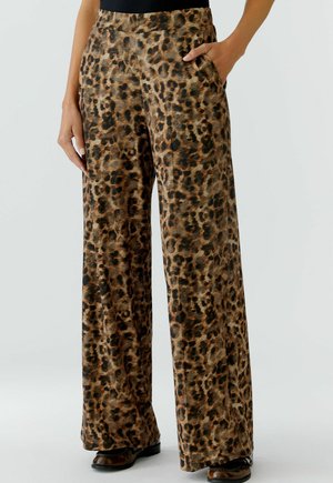 Vrouw in wijde broek met bruine en zwarte luipaardprint, met één hand in de zak, gecombineerd met donkere schoenen en een zwart shirt.