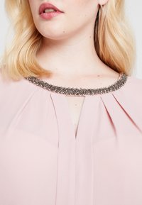 Ljusrosa blus med veck och en glittrande, utsmyckad halslinje med metalliska detaljer. Slät textur, enkel design.