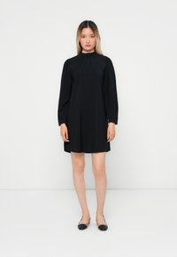 Abito nero con collo alto e maniche lunghe, caratterizzato da una vestibilità ampia e una texture liscia. Indossato con scarpe basse nere. Design minimalista.