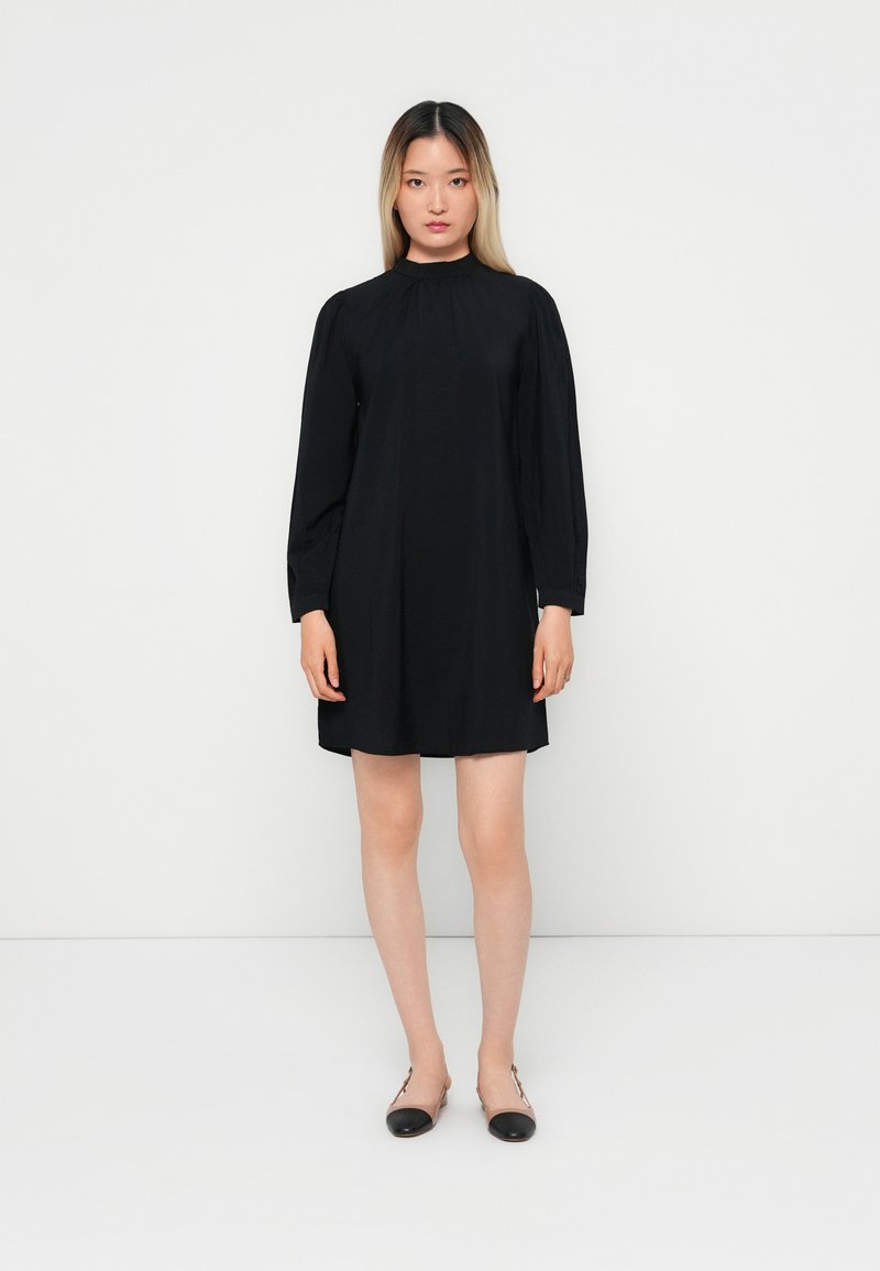 Abito nero con collo alto e maniche lunghe, caratterizzato da una vestibilità ampia e una texture liscia. Indossato con scarpe basse nere. Design minimalista.