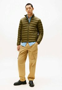 Vyras, apsirengęs alyvinių žalios spalvos siūtu švarku, šviesiai mėlynu marškinėliu, rusvomis krovinių kelnėmis ir juodais batais, stovi su rankomis kišenėse prieš baltą foną.