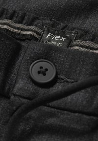 Gros plan de la ceinture d’un pantalon gris foncé montrant un bouton noir et une étiquette intérieure portant la mention « Flex On the go » avec une bordure élastique cousue.
