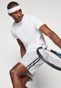 White athletic t-shirt with short sleeves, paired with gray shorts featuring green and blue stripes. Holding a tennis racket.