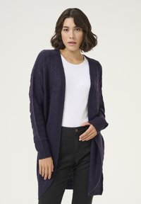 Marineblauer Strickcardigan mit offenem Vorderteil, langen Ärmeln und gerippten Bündchen. Geprägtes Muster überall, getragen über einem weißen Oberteil und dunklen Jeans.