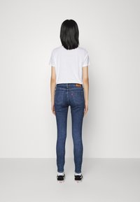 Vaqueros pitillo de mezclilla azul oscuro con cintura alta, bolsillos traseros sutiles y una etiqueta de cuero marrón. Combinados con una camiseta blanca y zapatillas negras.