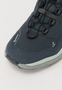 Námornícky modré turistické topánky s textúrovaným zvrškom, značkou GORE-TEX, elastickými šnúrkami a polstrovanou podrážkou s jemne zeleným akcentom.