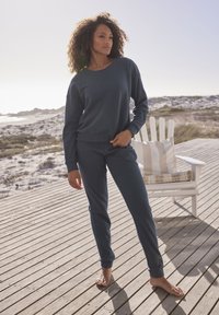 Dunkelblaues, geripptes Sweatshirt und passende Hose. Lockerer Schnitt, runder Halsausschnitt und strukturiertes Material. Barfuß auf einem Holzdeck, mit natürlichem Hintergrund.