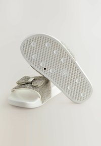 Baltas slīd sandals ar strukturētu zoli, kuriem ir mirdzošs sudraba loks uz siksniņas un gluda sintētiska virsma.