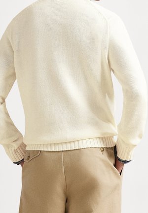 Osoba ubrana w kremowy sweter z dzianiny i beżowe spodnie, z rękami w kieszeniach, pokazana od tyłu na jasnym tle.