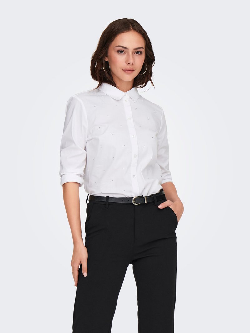Femme aux cheveux bruns mi-longs, debout, une main dans la poche, portant une chemise blanche à boutons et un pantalon noir avec ceinture.
