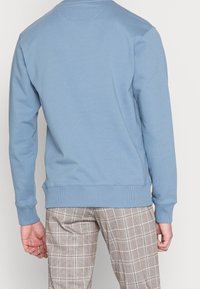 Světle modrá mikina s dlouhými rukávy, žebrovanými manžetami a lemem. Hladká textura a jednoduchý design; nošena s kostkovanými kalhotami.