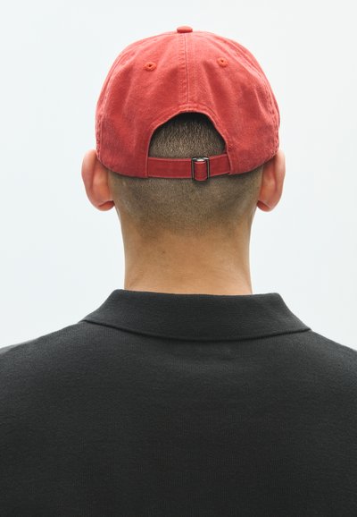 Ενήλικας που φοράει κόκκινο προσαρμοζόμενο καπέλο μπέιζμπολ και μαύρο πουκάμισο με γιακά, φαίνεται από πίσω σε απλό ανοιχτό φόντο.