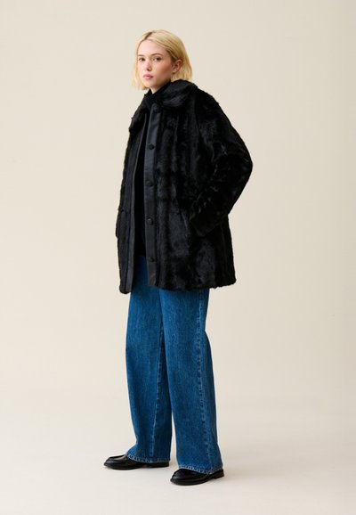 Cappotto in pelo sintetico nero con colletto e chiusura a bottoni, abbinato a jeans in denim blu a gamba larga e scarpe nere.