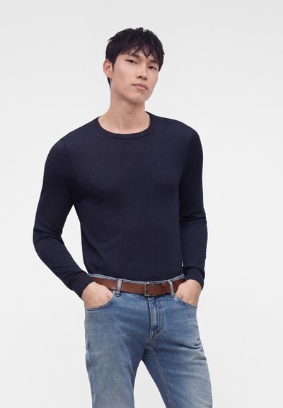 Homme portant une chemise ajustée bleu marine à manches longues, un jean bleu et une ceinture marron, debout les mains dans les poches devant un fond blanc uni.
