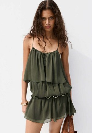 Femme aux longs cheveux bouclés porte une robe sans manches verte olive à plusieurs couches et tient un sac marron, sur un fond blanc uni.