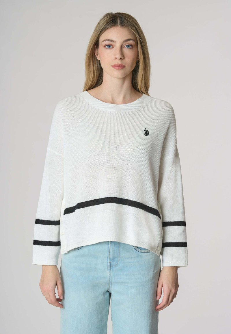 Maglione bianco a coste con strisce orizzontali nere, scollatura ampia e spalle ribassate. Presenta un piccolo emblema sul petto.