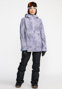 Chaqueta de nieve de color púrpura claro con diseño tie-dye, con un bolsillo en el frente y media cremallera, combinada con pantalones de nieve negros y guantes negros, acompañado de un gorro y gafas de protección.
