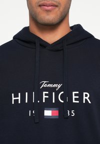 Granatowa bluza z kapturem z białym napisem "Tommy HILFIGER" oraz małym akcentem w postaci logo w formie flagi. Materiał wygląda na miękki, kaptur z regulowanym sznurkiem.