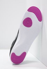 Suola di una scarpa Nike nera, con una parte inferiore bianca strutturata e un distintivo pad in gomma magenta. La scarpa è appoggiata su una superficie bianca.