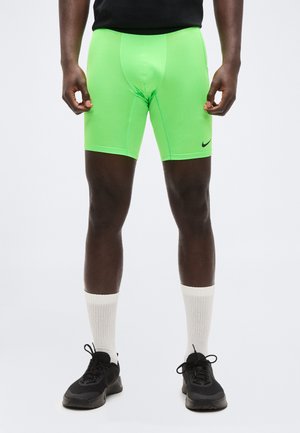 Homme portant un short de compression Nike vert vif, des baskets noires, des chaussettes blanches montantes, debout devant un fond uni.