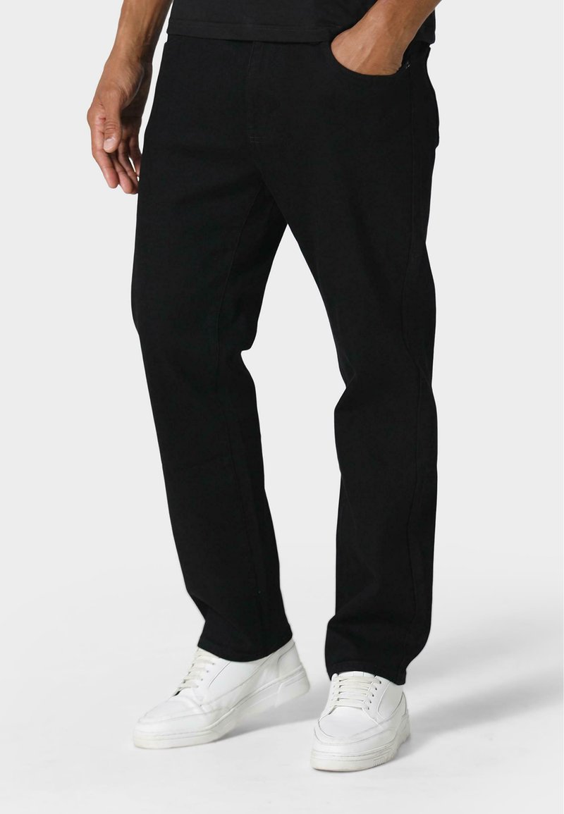 Pantalones de mezclilla negros con corte recto, con bolsillos laterales y un ligero estiramiento. Llevados con zapatillas blancas, sobre un fondo neutro.