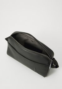 Pochette en cuir noir avec fermeture éclair, de forme plate, texture lisse, arborant un logo en relief subtil et un intérieur en tissu doux.