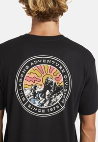T-shirt nera in cotone con una grafica circolare che presenta montagne, un fiume e un sole stilizzato nei colori rosa, giallo e blu. Il testo include "Billabong Adventure Division" e "Dal 1973".