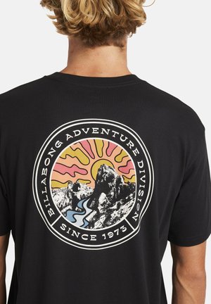 Black cotton t-shirt featuring a circular graphic with mountains, a river, and a sunburst in pink, yellow, and blue. Text includes "Billabong Adventure Division" and "Since 1973."