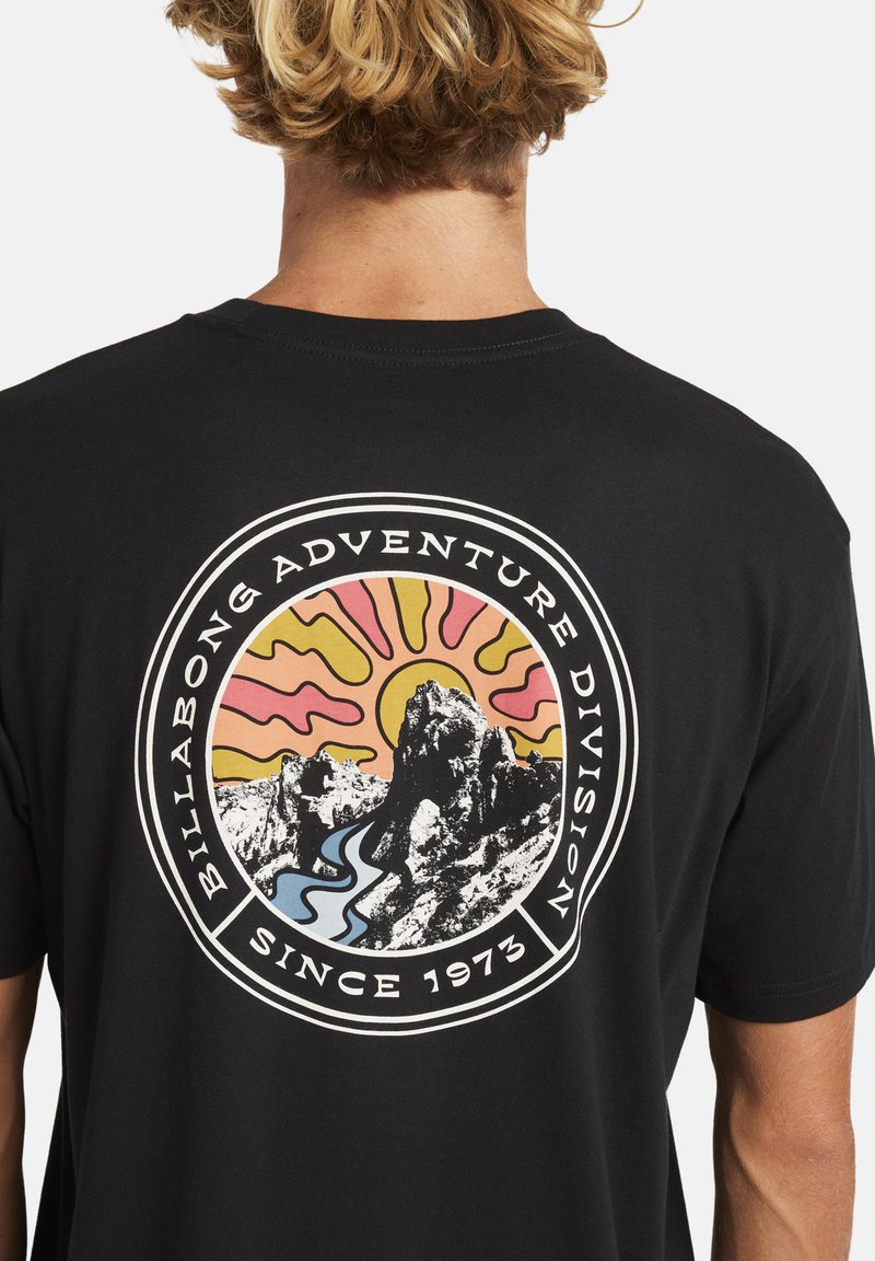 T-shirt nera in cotone con una grafica circolare che presenta montagne, un fiume e un sole stilizzato nei colori rosa, giallo e blu. Il testo include "Billabong Adventure Division" e "Dal 1973".