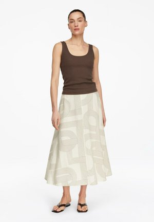 Kobieta ubrana w brązowy top bez rękawów, kremową spódnicę w literę A z geometrycznymi wzorami oraz czarne sandały na otwartym palcu, stojąca na tle białego tła.