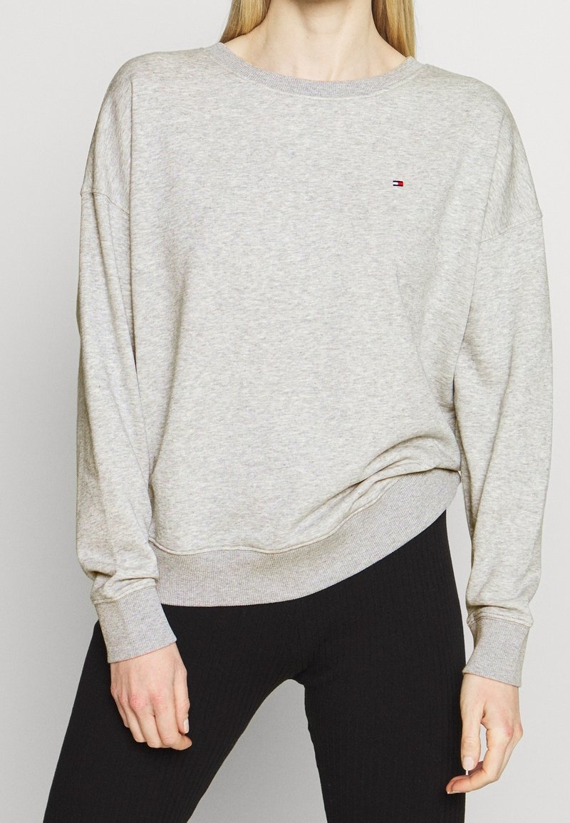 Femme portant un sweat-shirt à manches longues gris clair avec un petit logo Tommy Hilfiger et un pantalon noir ajusté, debout devant un fond uni.