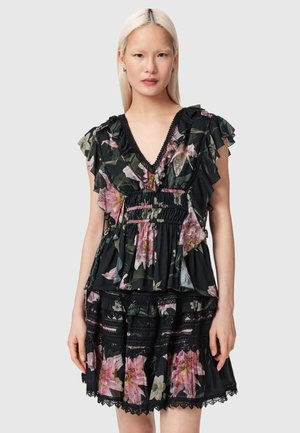 Vrouw in een zwarte jurk met bloemenprint en ruches, met roze en groene bloemen, met een diepe V-hals en kanten details op de rok.