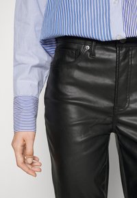 Černé kožené kalhoty s hladkou texturou, vybavené dvěma předními kapsami a kontrastním prošíváním. Oblečení dopl�ňuje modro-bílá pruhovaná košile.