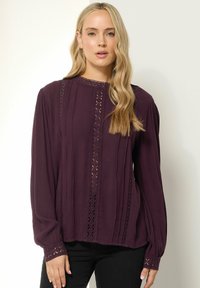 Blusa a maniche lunghe di un profondo colore viola, realizzata in tessuto leggero, con pieghe verticali, dettagli in pizzo e polsini abbottonati.