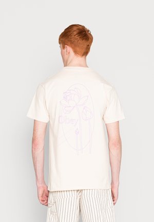 Person mit roten Haaren, die ein cremefarbenes T-Shirt mit einem hellvioletten Blumenmuster und dem Text "Obey" auf dem Rücken sowie gestreifte hellfarbige Hosen trägt.