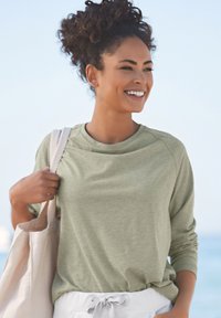 Langärmliges, hellgrünes Baumwollhemd mit Rundhalsausschnitt und Raglanärmeln, kombiniert mit weißen Jogginghosen mit Kordelzug und einer Tragetasche aus Leinen.