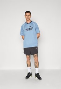 Lys blå kortærmet trøje med sort Puma-logo, marineblå krave, parret med sorte shorts og sorte sneakers med hvide og grønne accenter.