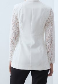Biały żakiet z długimi koronkowymi rękawami, zdobiony skomplikowanymi wzorami florystycznymi. Gładki materiał o strukturalnym kroju i dopasowanej sylwetce.