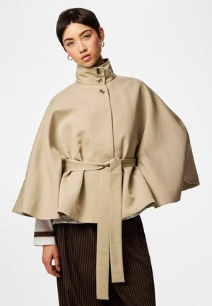 Žena v béžovom plášti s opaskom a vysokým golierom s tromi gombíkmi, v kombinácii s tmavými nohavicami s tenkými pásikmi a kruhovými náušnicami.