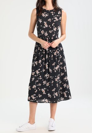 Sort ærmeløs kjole med blomstermønster i nuancer af hvid og fersken, med en tætsiddende overdel og sammenrevne talje, parret med hvide sneakers.