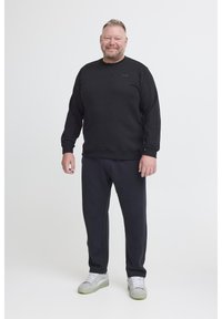 Un uomo sorride indossando un felpa nera, pantaloni neri e sneakers grigie chiare su uno sfondo bianco semplice.